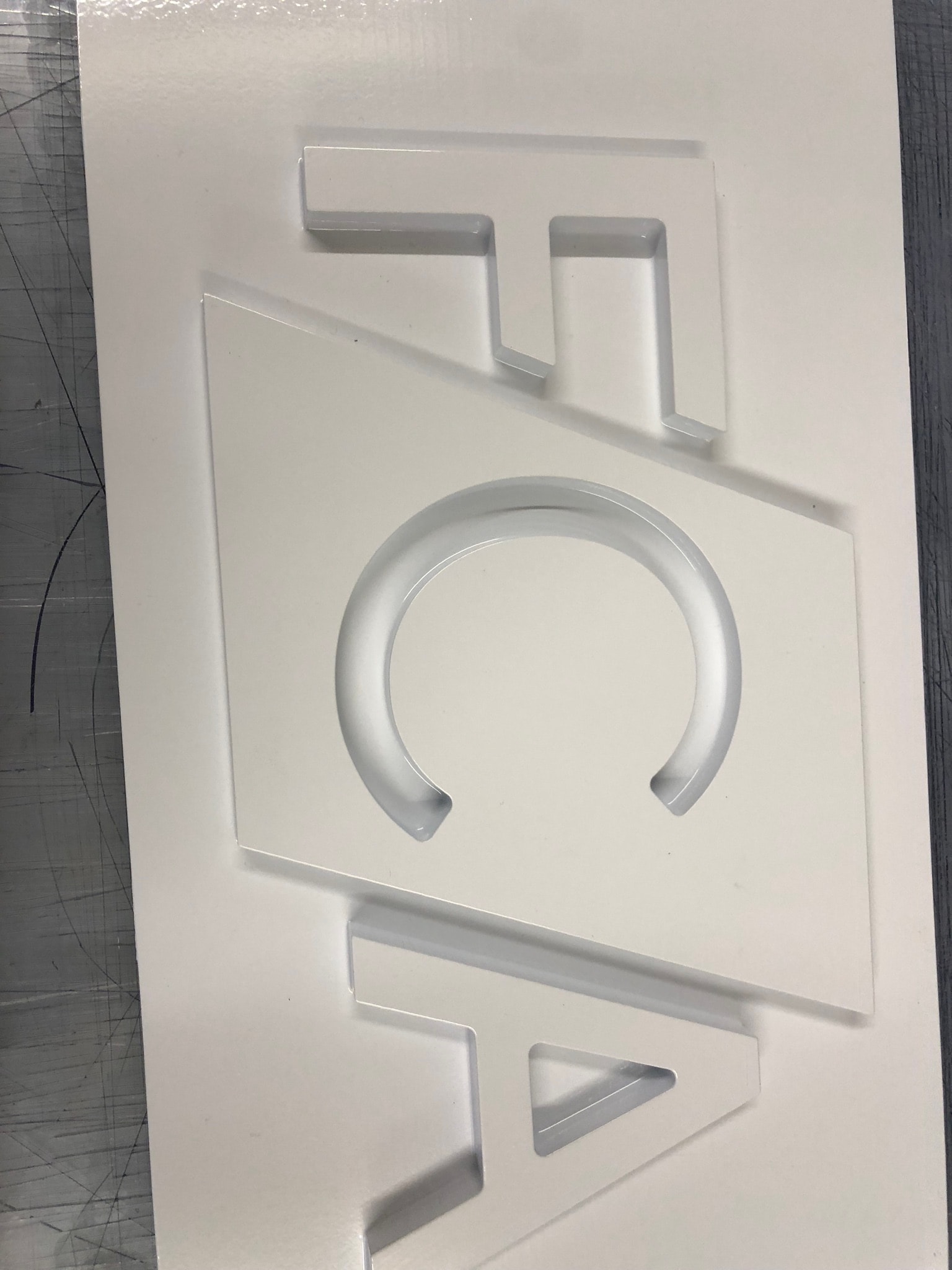 White acrylic sheet with precision CNC-routed recessed pockets and letter-like shapes on a workshop bench, indicating flat-bed milling work-in-progress.