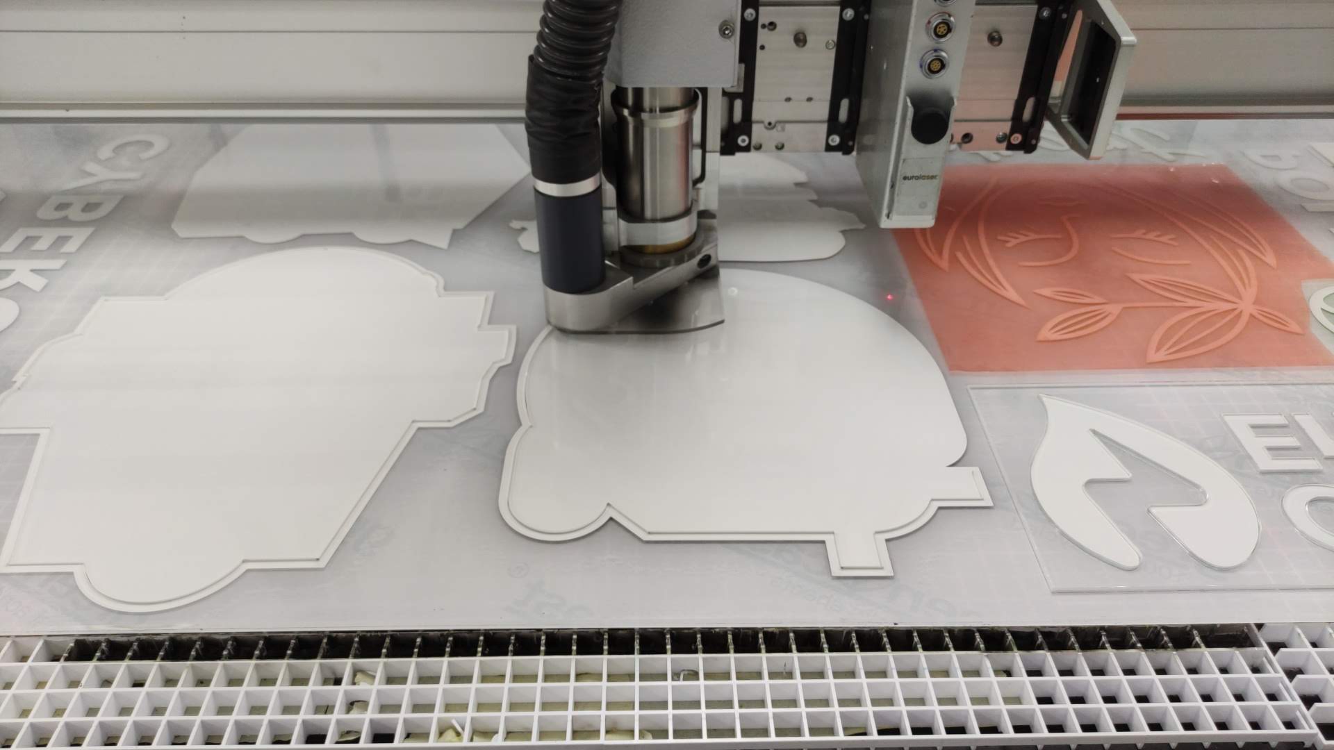 CNC router cutting white acrylic sign shapes on a flatbed table, with extraction hose and nested parts visible in a fabrication workshop.