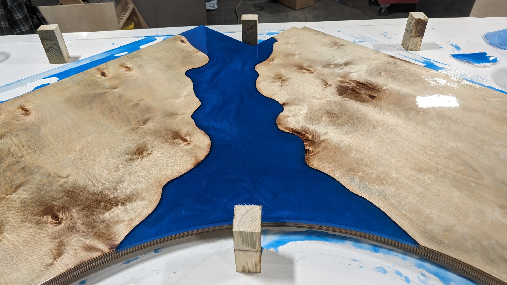 Top-down workshop photo of a river table in progress showing two plywood slabs joined with a blue epoxy resin fill, held with wooden blocks during curing.