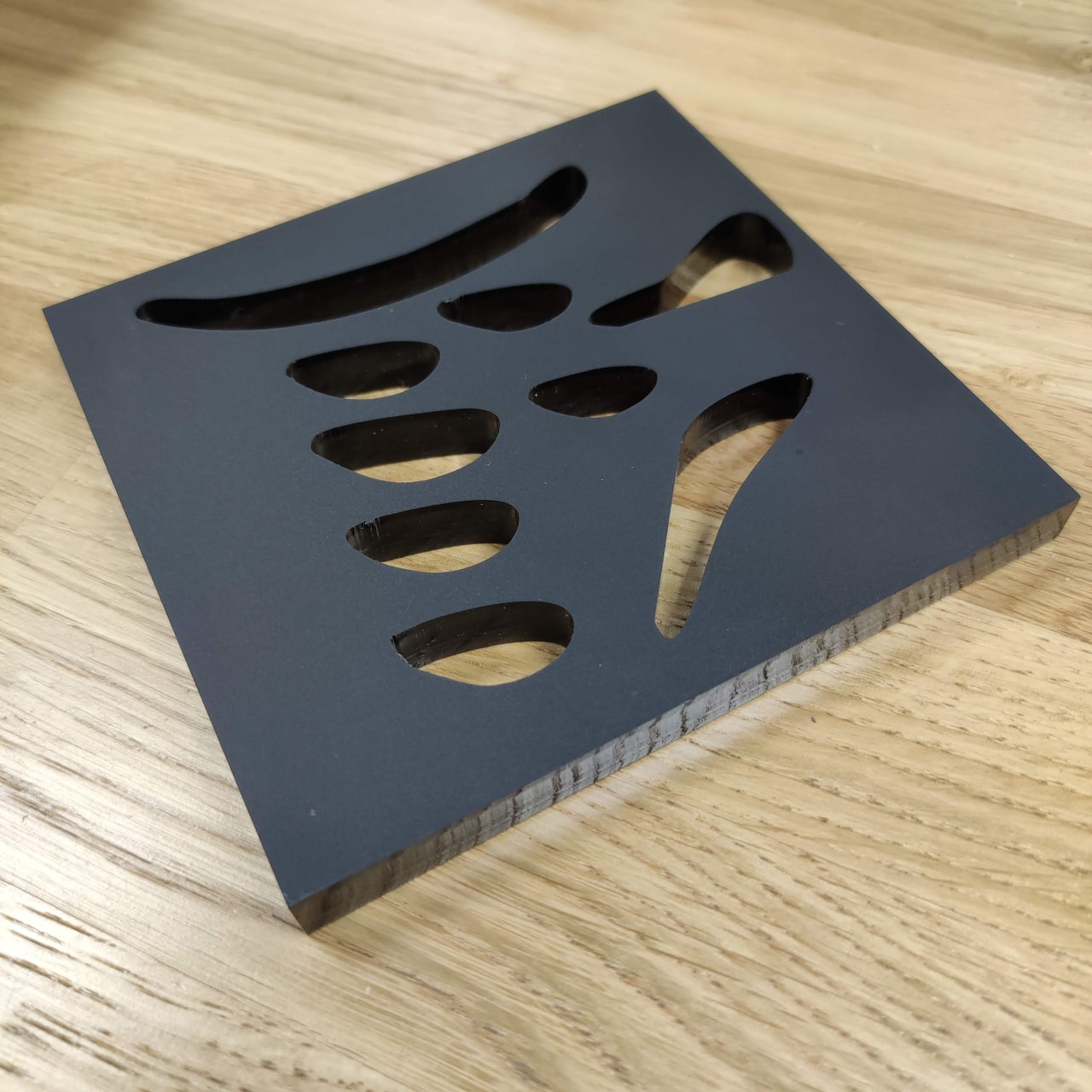 Black coated routed panel with organic teardrop cutouts photographed on a wooden workbench; a CNC-machined sample or component.