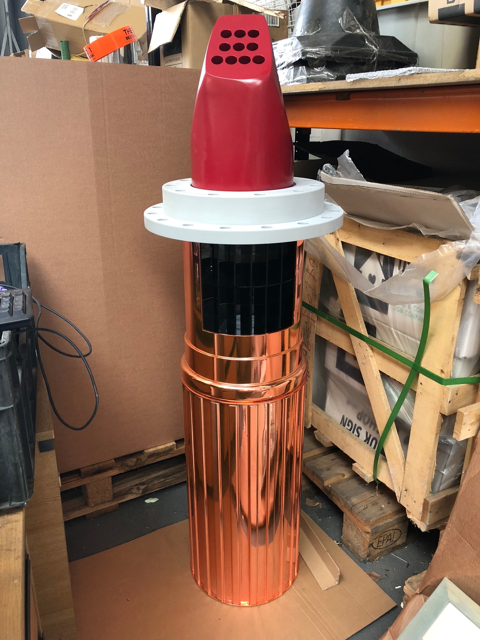 Polished copper cylindrical fabricated component with a red vented top and flange photographed in a workshop among pallets and crates.