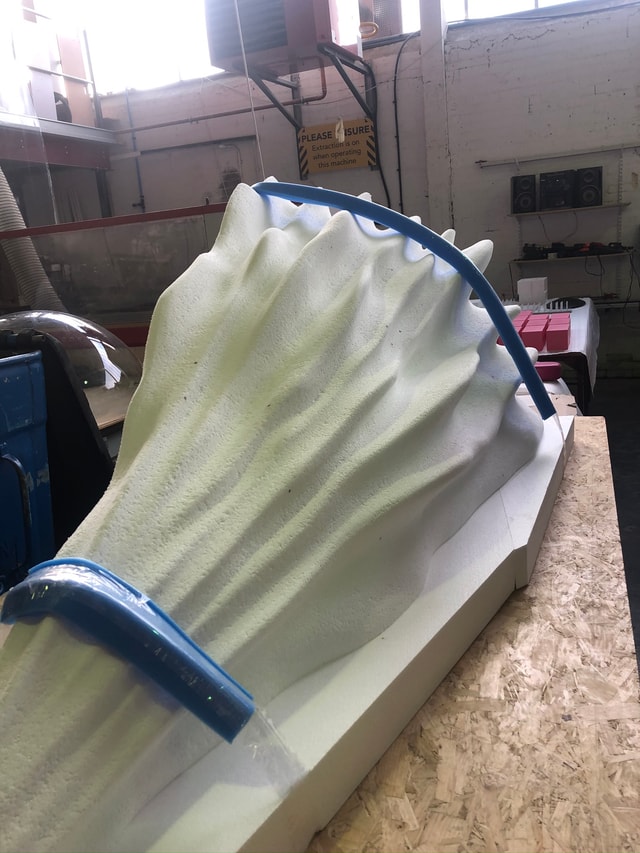 Large carved foam sculpture or mould on a workshop bench, photographed in a fabrication workshop showing tooling and supports.