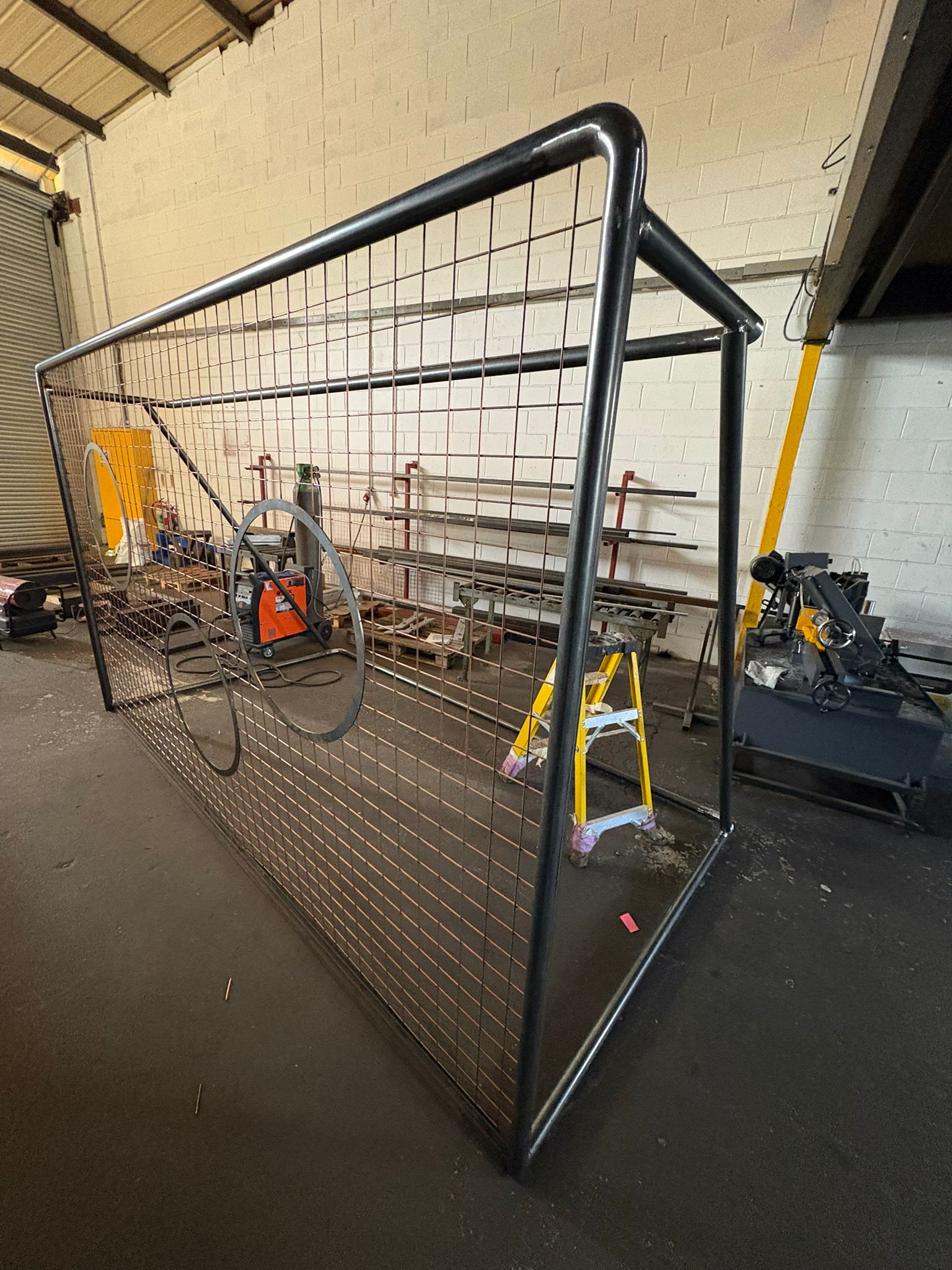 Welded tubular steel frame with wire mesh and circular cutouts pictured in a fabrication workshop — metalwork in progress suitable for partitions or display structures.