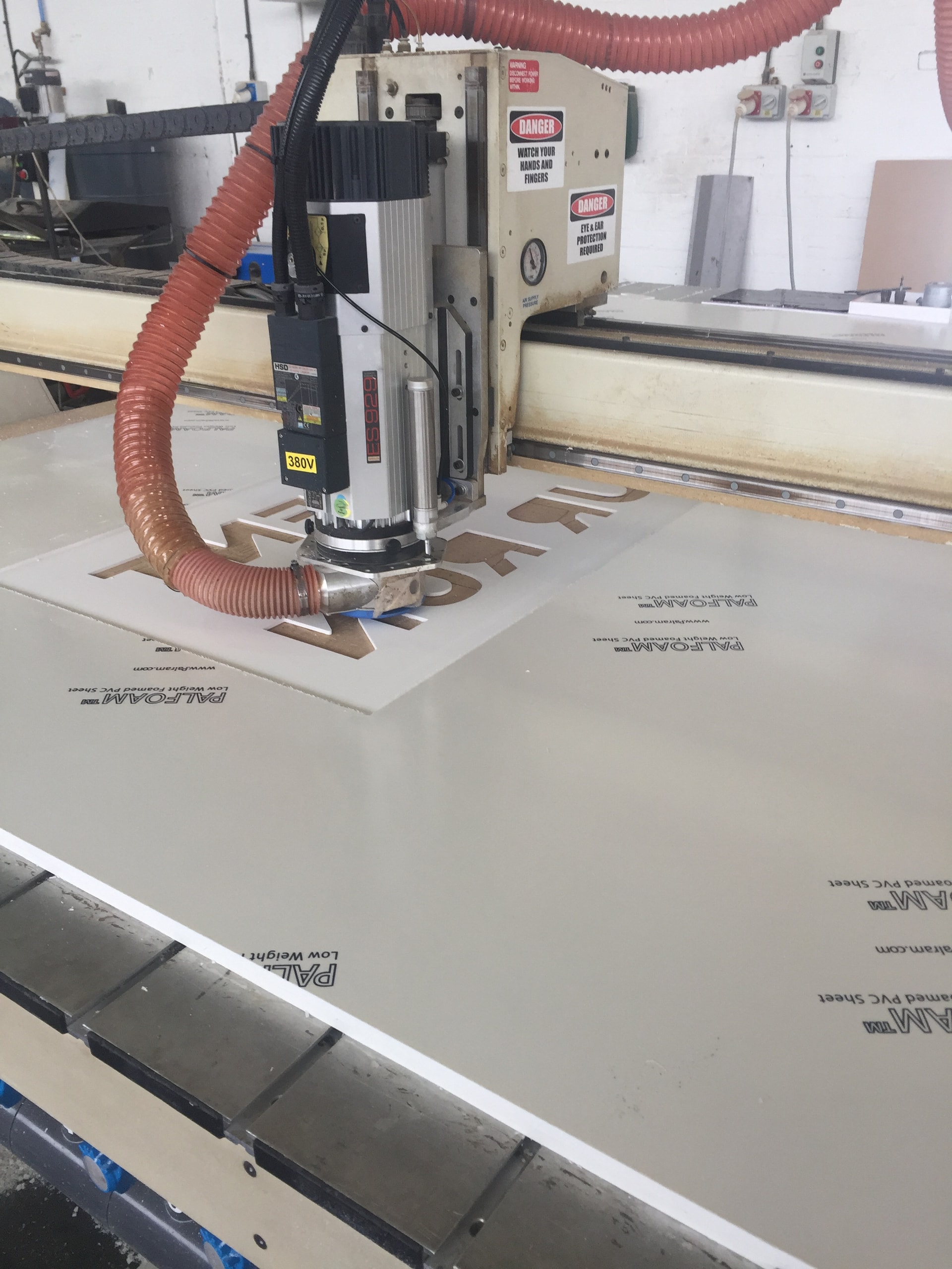 CNC router cutting letter shapes from a PALFOAM sheet on a flat-bed table with dust extraction in a fabrication workshop.