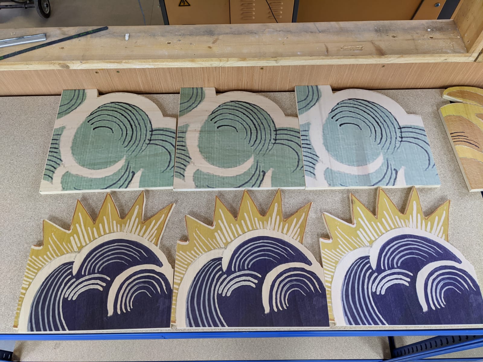 CNC-cut plywood panels with printed wave-and-sun graphics laid out on a workshop bench, showing finished cut shapes ready for assembly or installation.