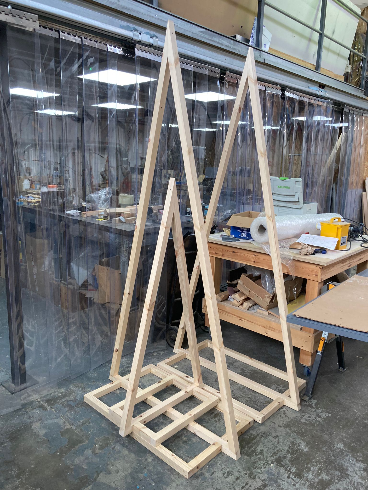 Wooden A-frame display stands being fabricated in a workshop, shown with benches, tools and materials in the background.