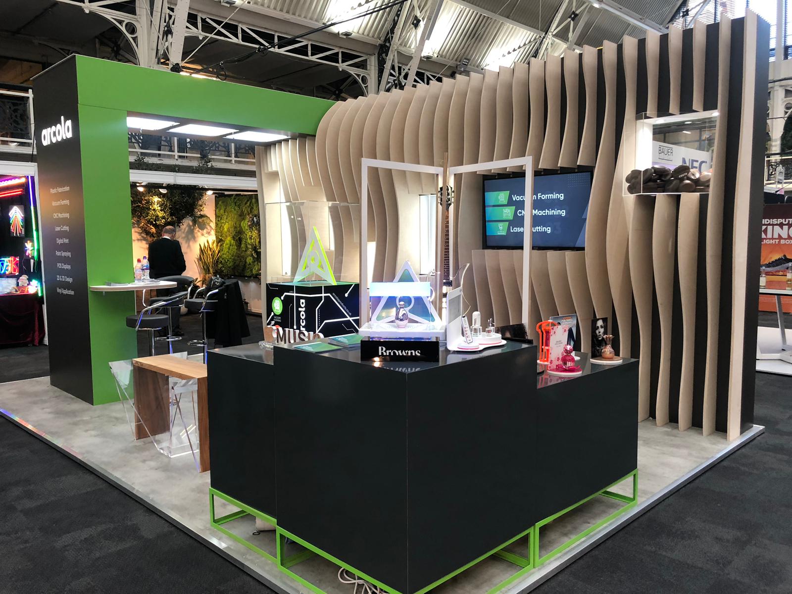 Arcola exhibition stand at a trade show with black counters, curved timber slat backdrop, illuminated demo displays and prominent Arcola branding.