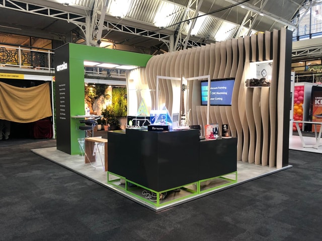 Arcola exhibition stand at a trade show featuring a green arch with illuminated logo, wave-like wooden backdrop, display counters and a screen highlighting fabrication services.