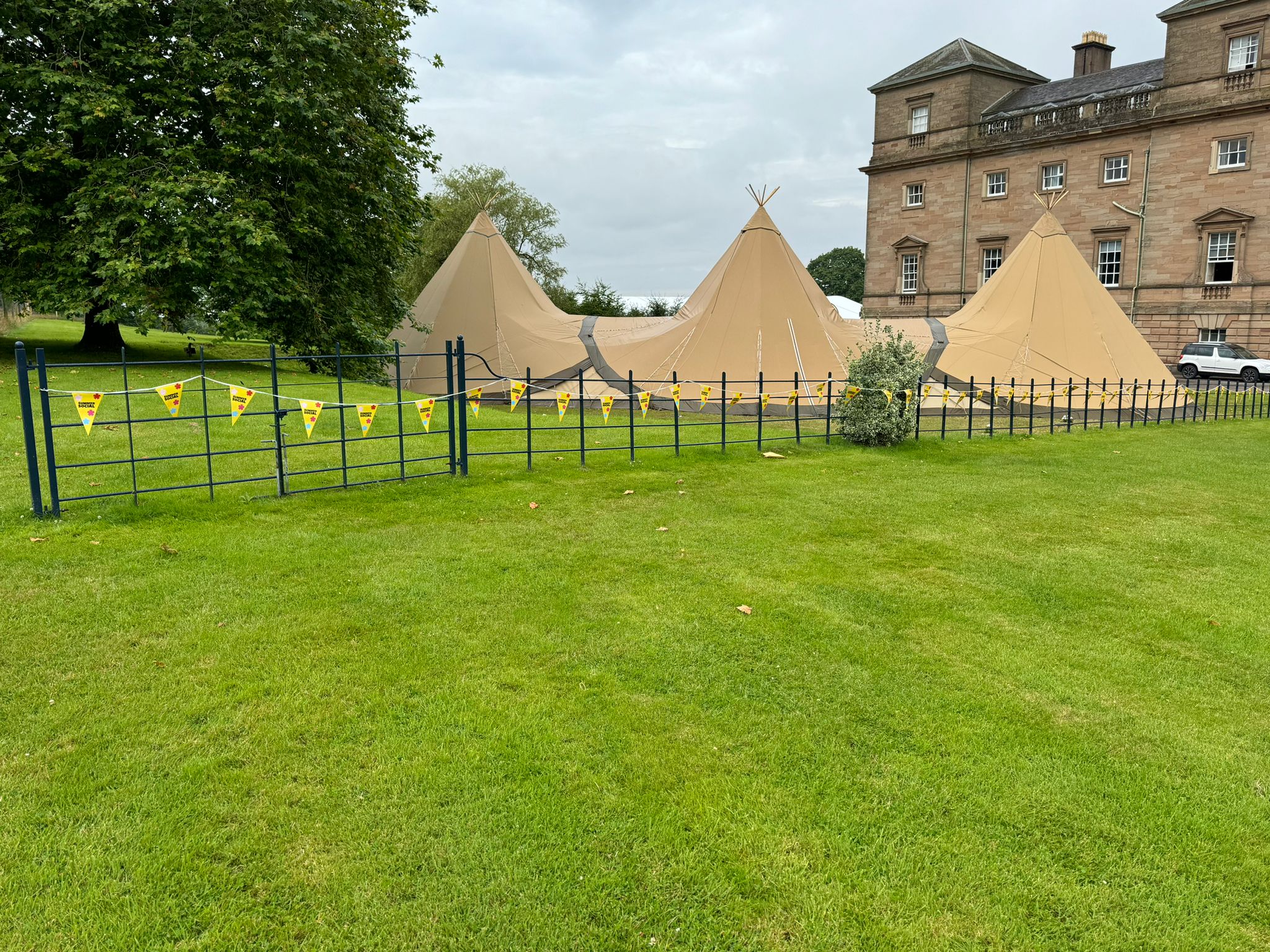 Outdoor event setup with large bell tents and branded bunting on temporary fencing in front of a historic building, photographed on a grassy lawn.