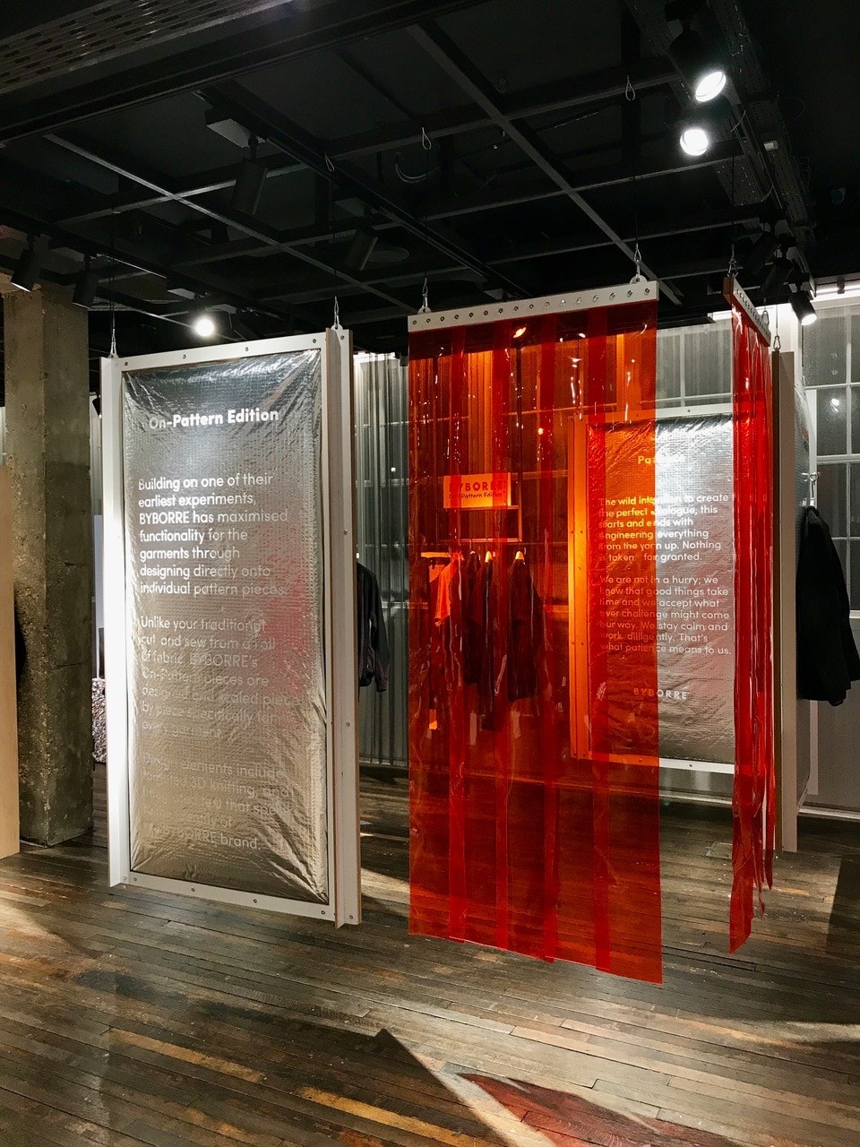 Interior exhibition/pop-up retail installation with framed information panels and red translucent strip curtains showcasing garments in a curated event space.