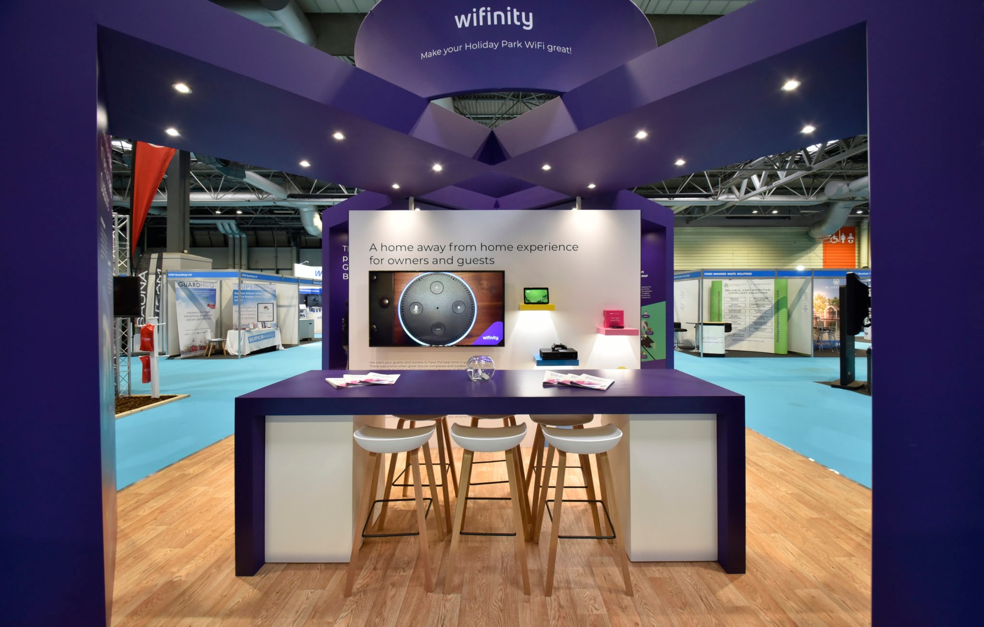 Branded purple Wifinity exhibition stand at a trade show, featuring a central counter, stools, display screen and illuminated overhead signage.