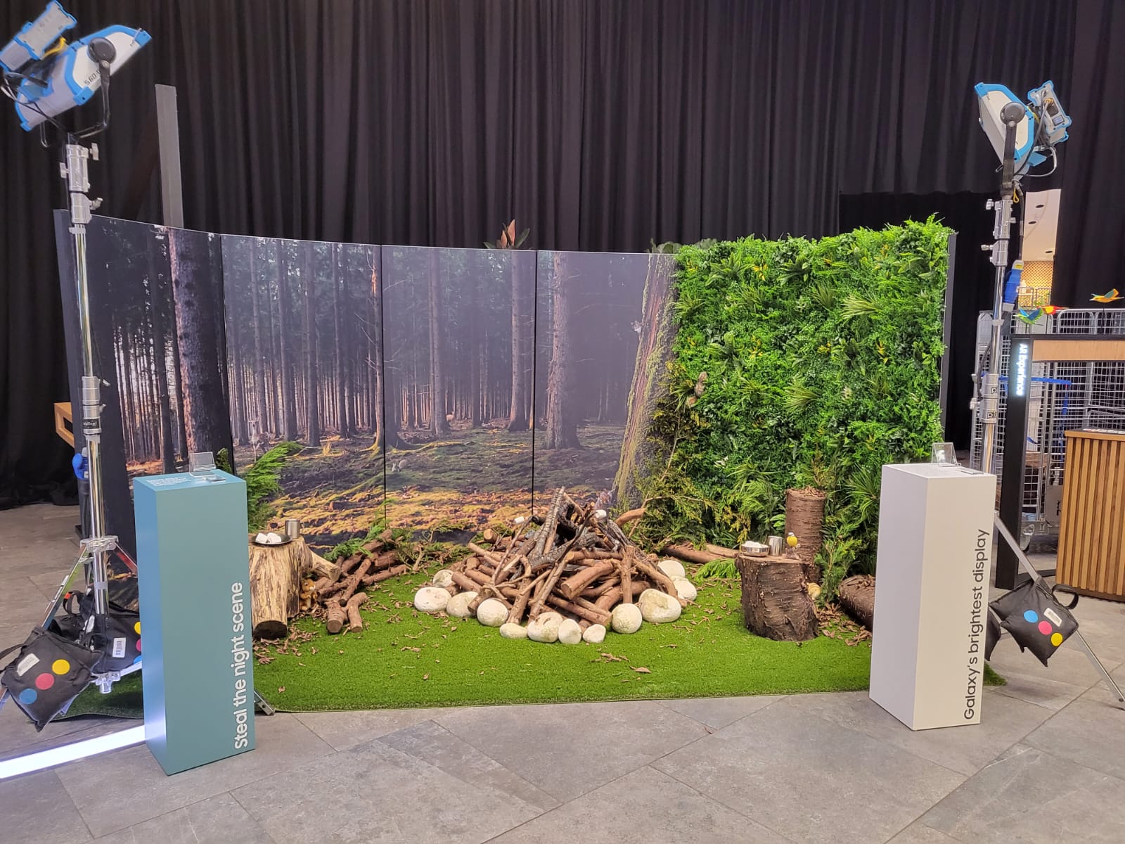 Exhibition stand with a large printed forest backdrop, living green wall, artificial turf and natural props, plus branded demo pedestals for a product display.