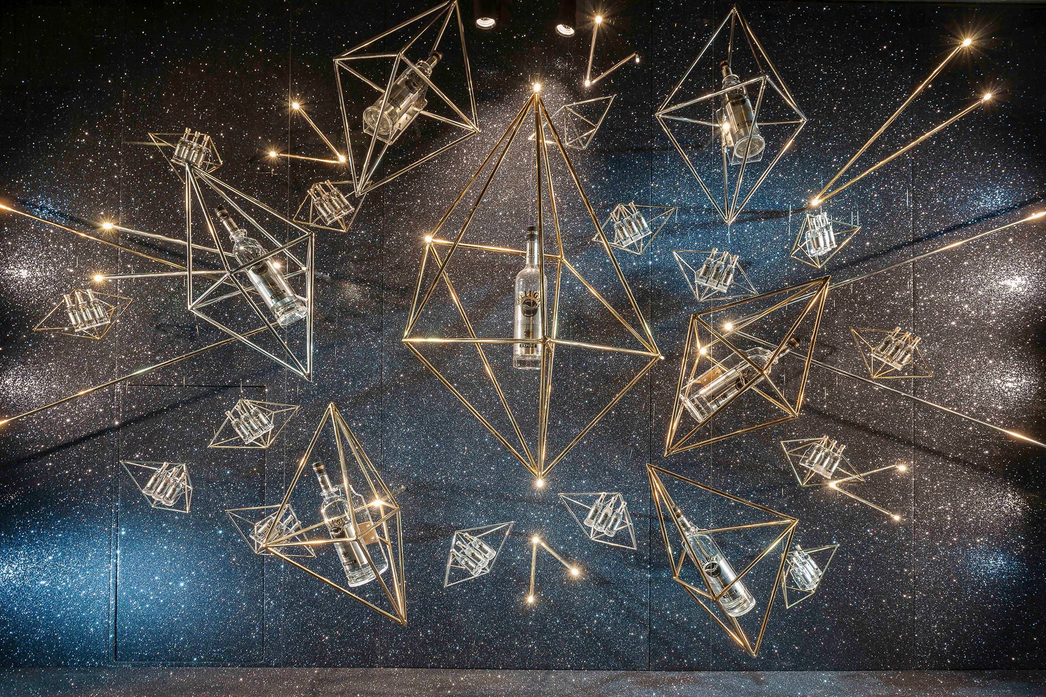 Suspended geometric brass frames holding glass bottles with integrated lighting against a starry glitter wall — an experiential retail/event installation and design concept.