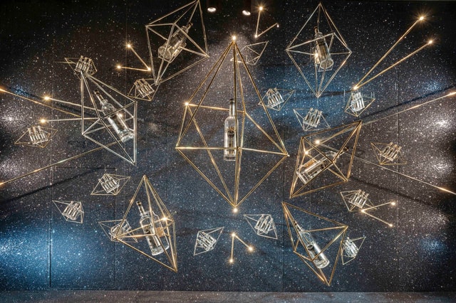 Suspended geometric brass frames holding glass bottles with integrated lighting against a starry glitter wall — an experiential retail/event installation and design concept.