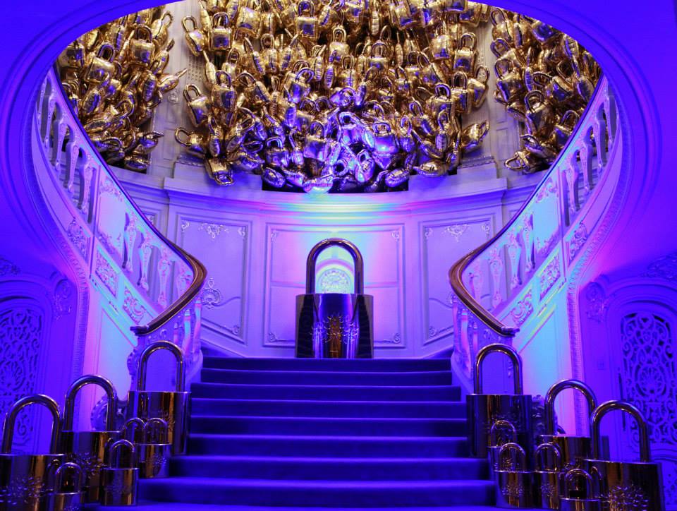Dramatic indoor event installation of oversized gold padlocks arranged on a grand staircase with purple and blue lighting.