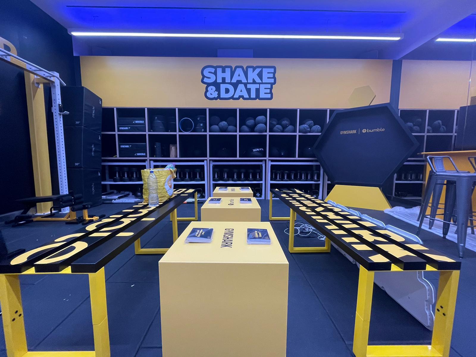 Branded Gymshark experiential activation 'Shake & Date' with yellow display plinths, benches and hexagonal backdrop in an indoor pop-up/event space.
