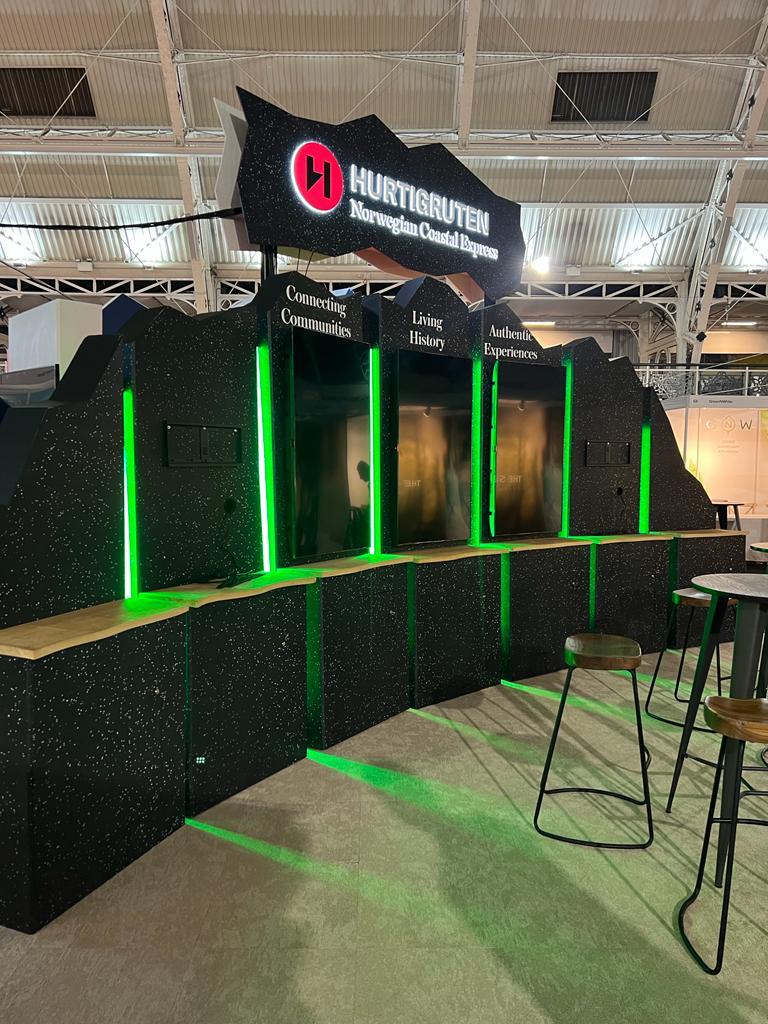 Finished exhibition booth for Hurtigruten featuring illuminated brand signage and vertical green LED accents, installed in a trade-show environment.