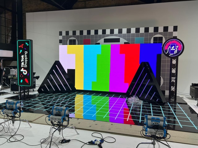 Studio event stage featuring a large LED video wall, branded vertical TikTok display and circular neon sign on truss, with reflective grid floor and studio lighting.