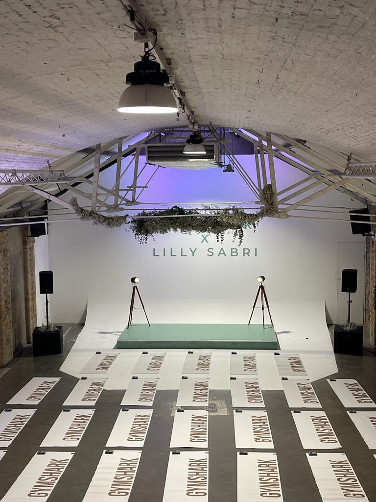 Event stage and branded backdrop for Lilly Sabri with repeated Gymshark floor graphics in an industrial event space; stage lighting and speakers visible.