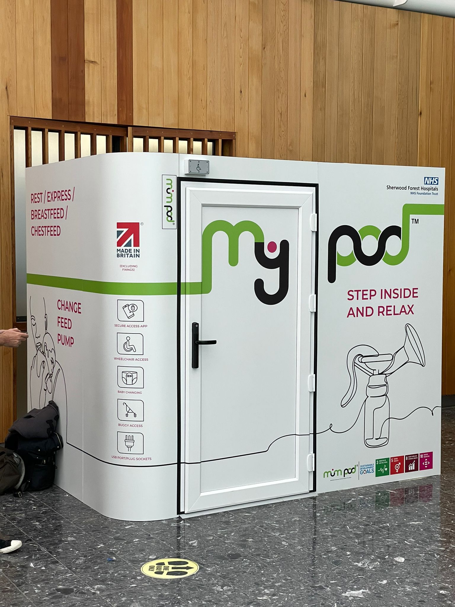 Freestanding MyPod nursing and rest pod installed indoors with full-coverage printed graphics, accessibility icons and branded signage, offering a private space for feeding and relaxation.