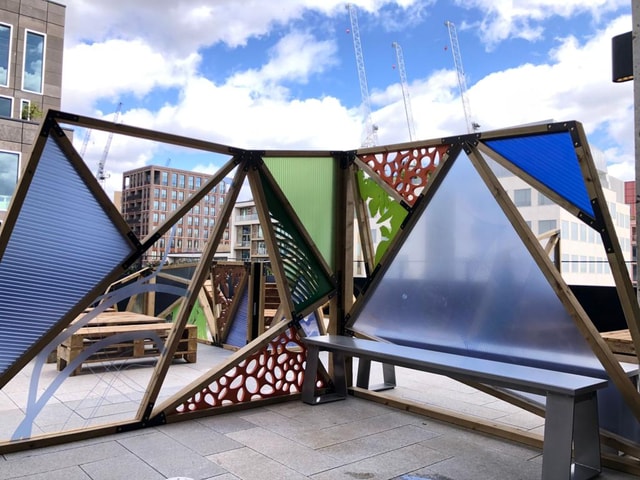 Temporary outdoor pavilion with triangular timber frames and coloured polycarbonate panels forming seating and partitions, installed in an urban plaza for an event or pop-up space.