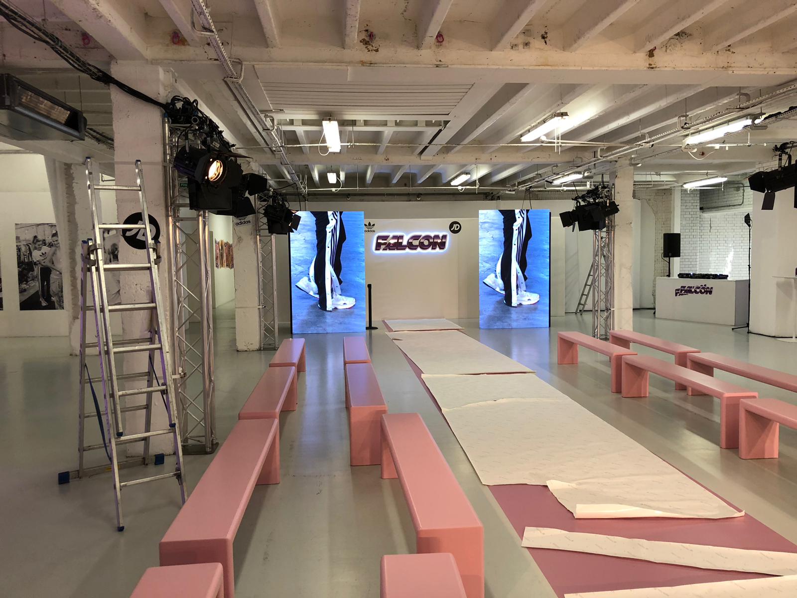 Interior event space staged for a fashion runway with pink benches, a central catwalk and LED screens flanked by truss lighting.