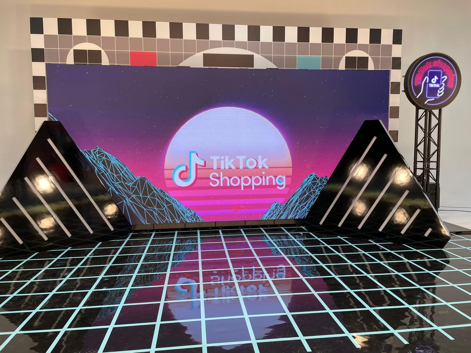 Branded event stage with a large LED screen showing the TikTok Shopping logo, reflective grid floor and glossy triangular set pieces in an experiential installation.