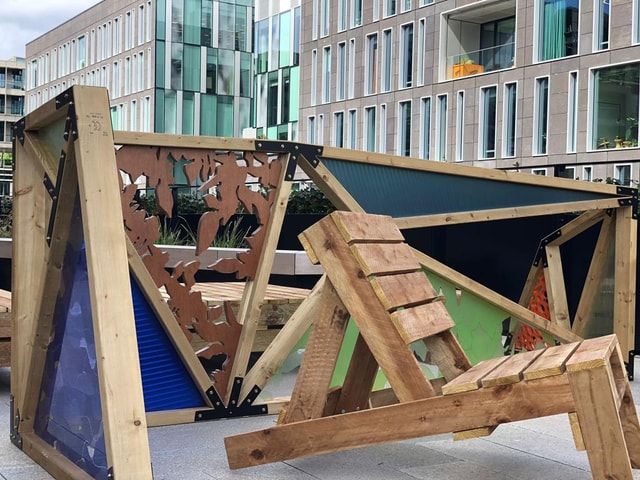 Modular timber seating with decorative laser-cut panels and colored corrugated infill installed in an urban plaza in front of office buildings.