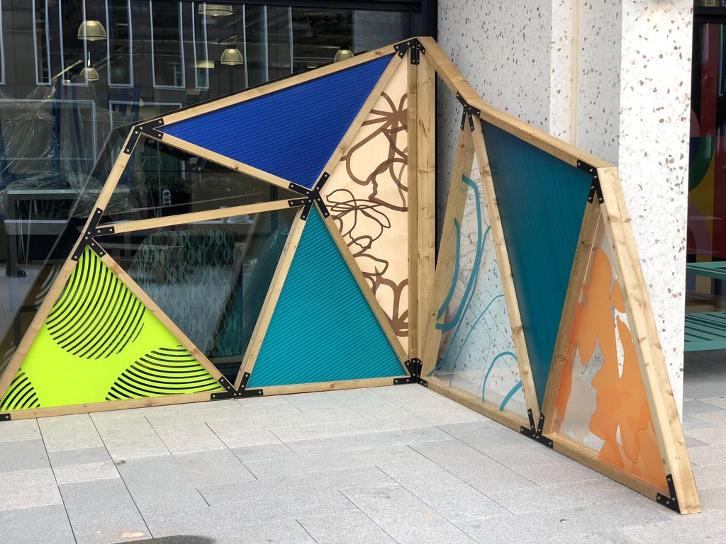 Freestanding triangular timber-frame installation with colourful large-format printed panels forming a decorative outdoor partition in front of a glass-fronted building.