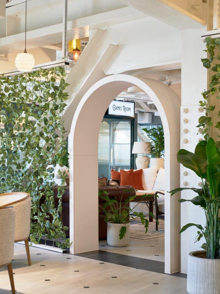 Bright modern office lounge with a decorative archway leading to a 'Games Room', featuring plants, comfortable seating and clear internal wayfinding signage.