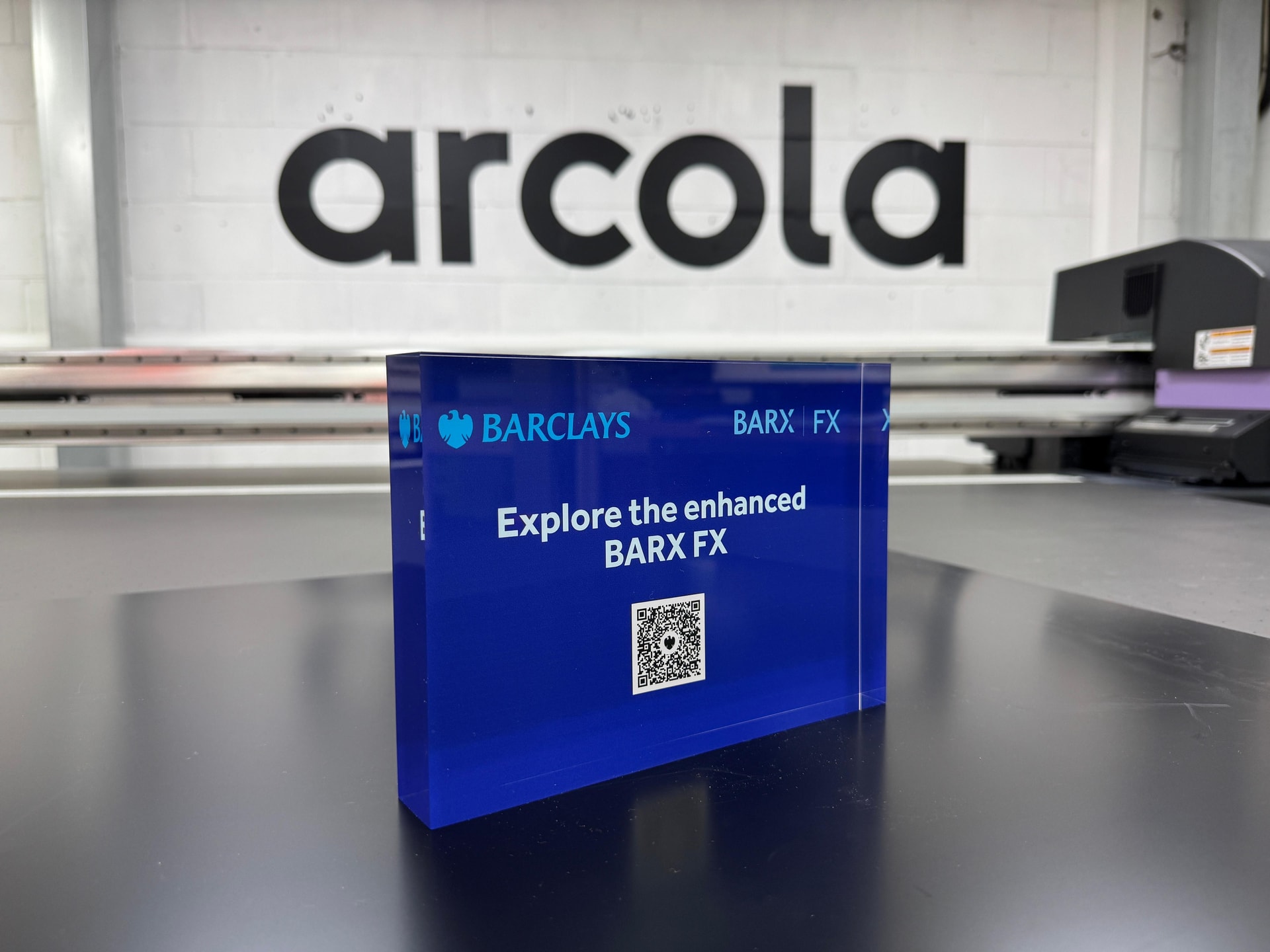 Blue acrylic promotional block with Barclays branding and QR code photographed on a workshop bench in front of a UV flatbed printer and Arcola branded wall.