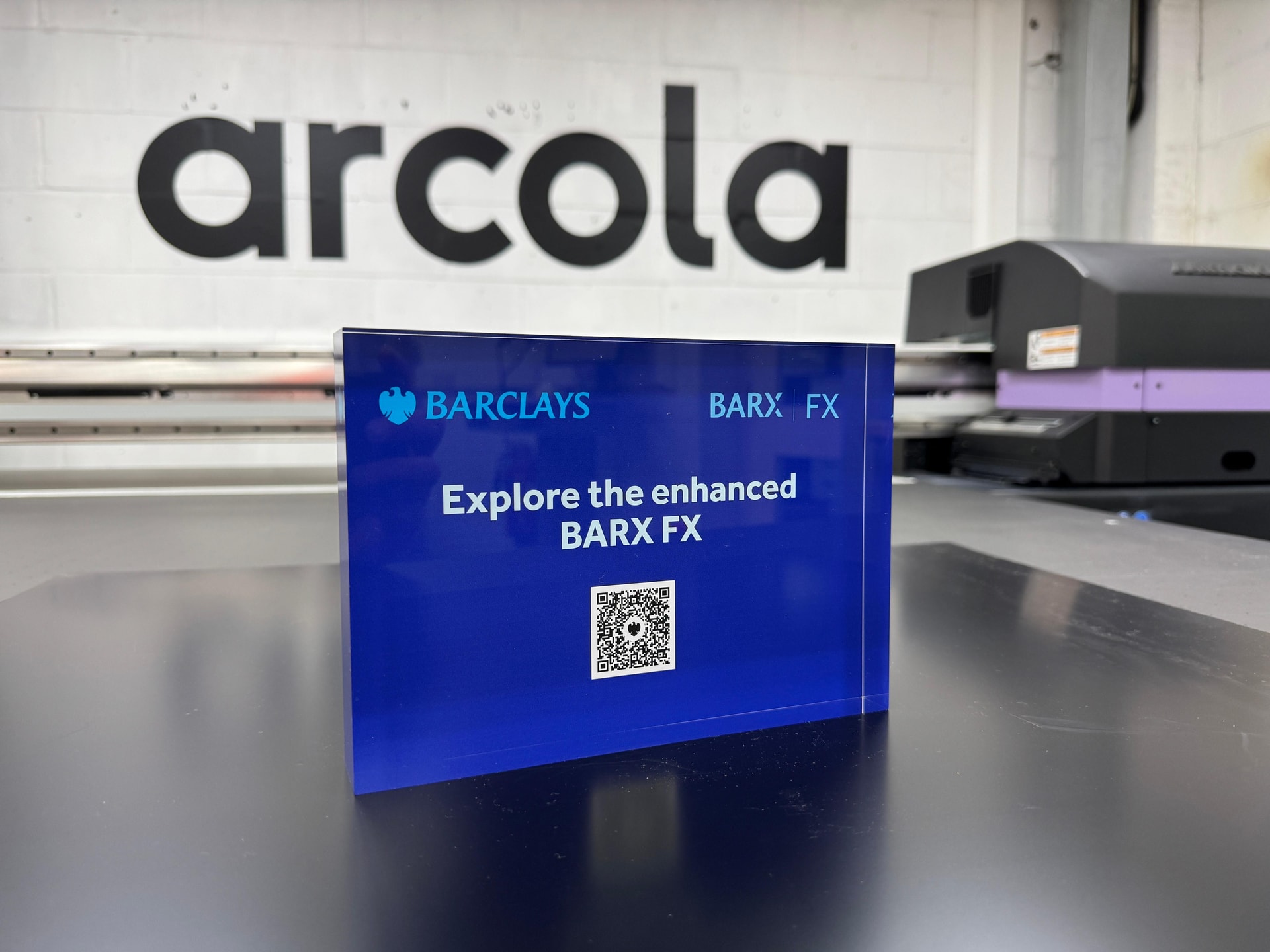 Blue acrylic counter-top promotional plaque printed with Barclays branding and a QR code, photographed on a workshop bench with a large-format printer and Arcola logo in the background.