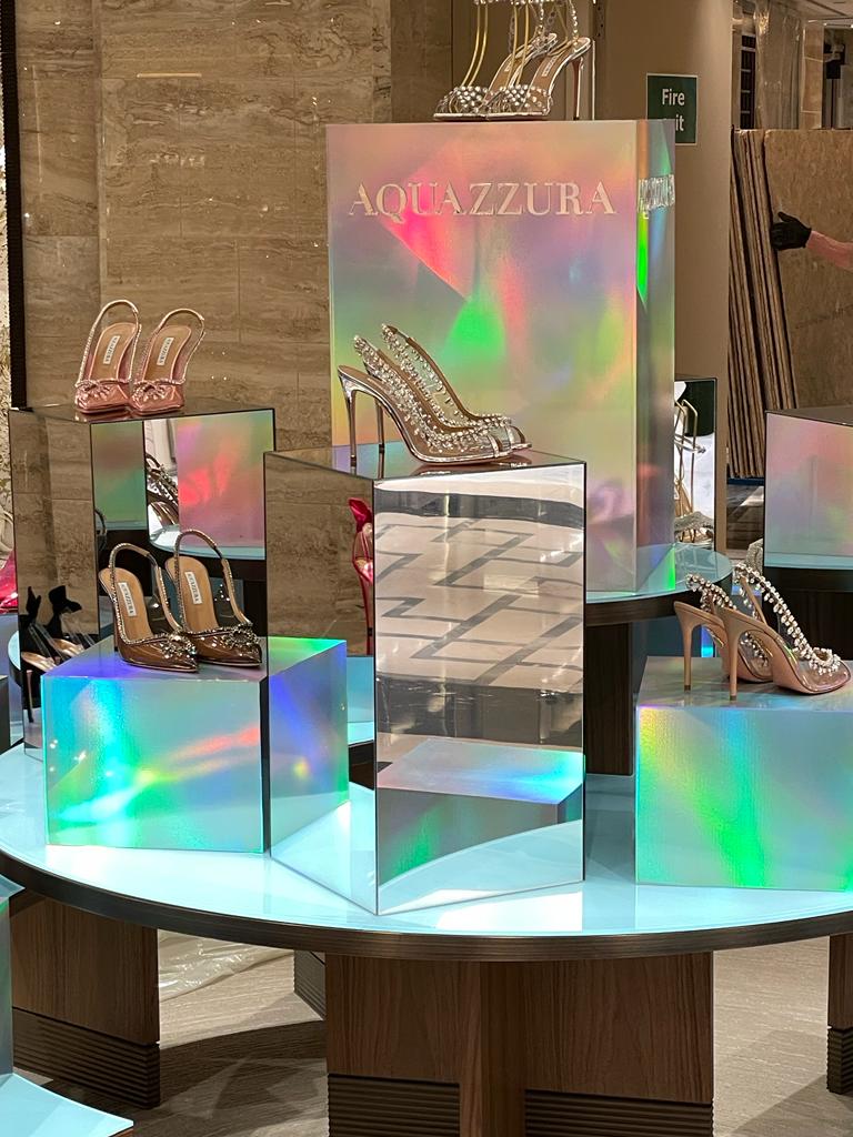 Iridescent mirrored display pedestals showcasing Aquazzura high-heeled shoes on a circular retail table, highlighting holographic acrylic fixtures and reflective surfaces for a luxury store display.