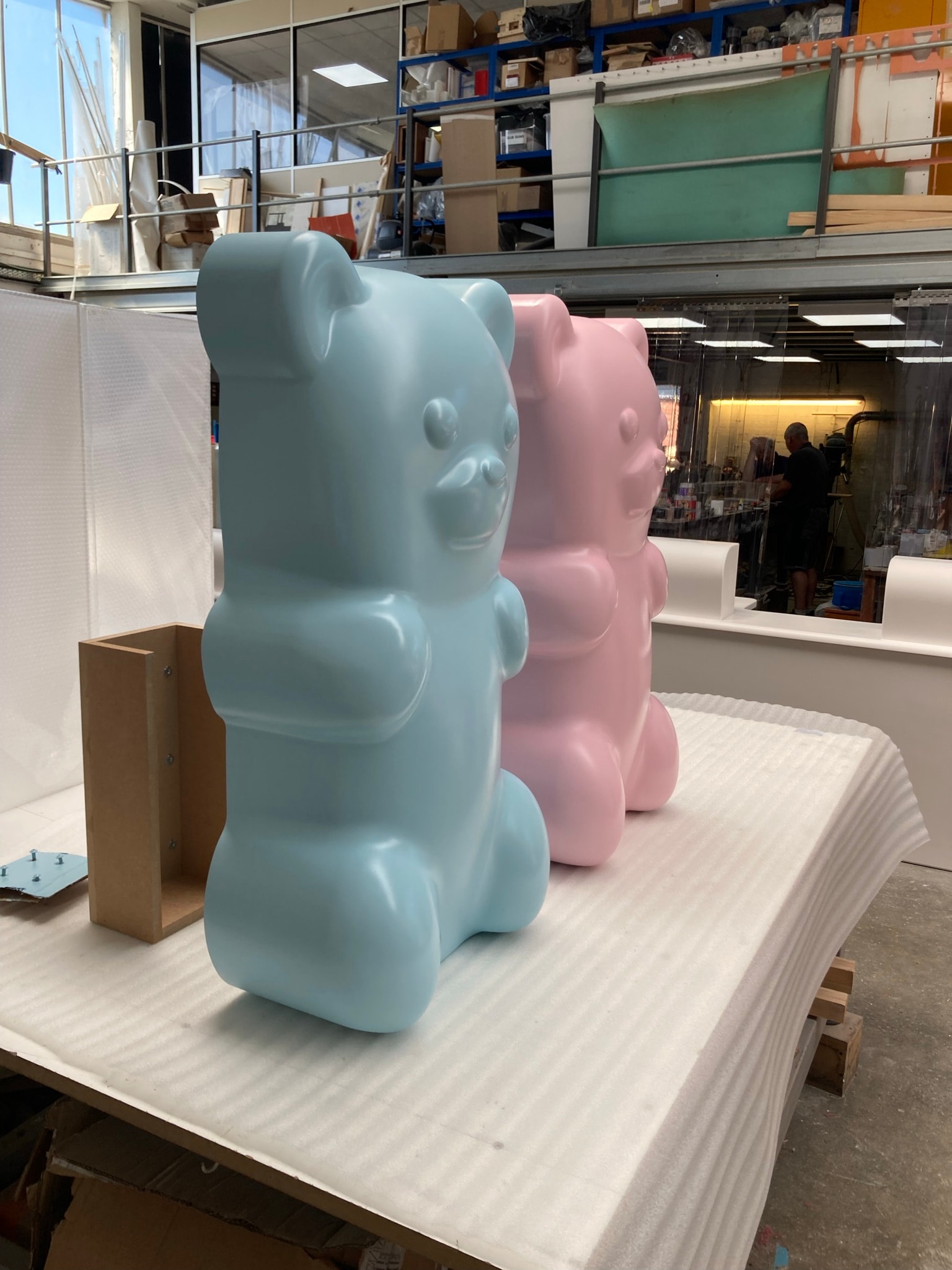 Pastel blue and pink molded bear display figures with glossy finish on a foam-covered workbench in a fabrication workshop.