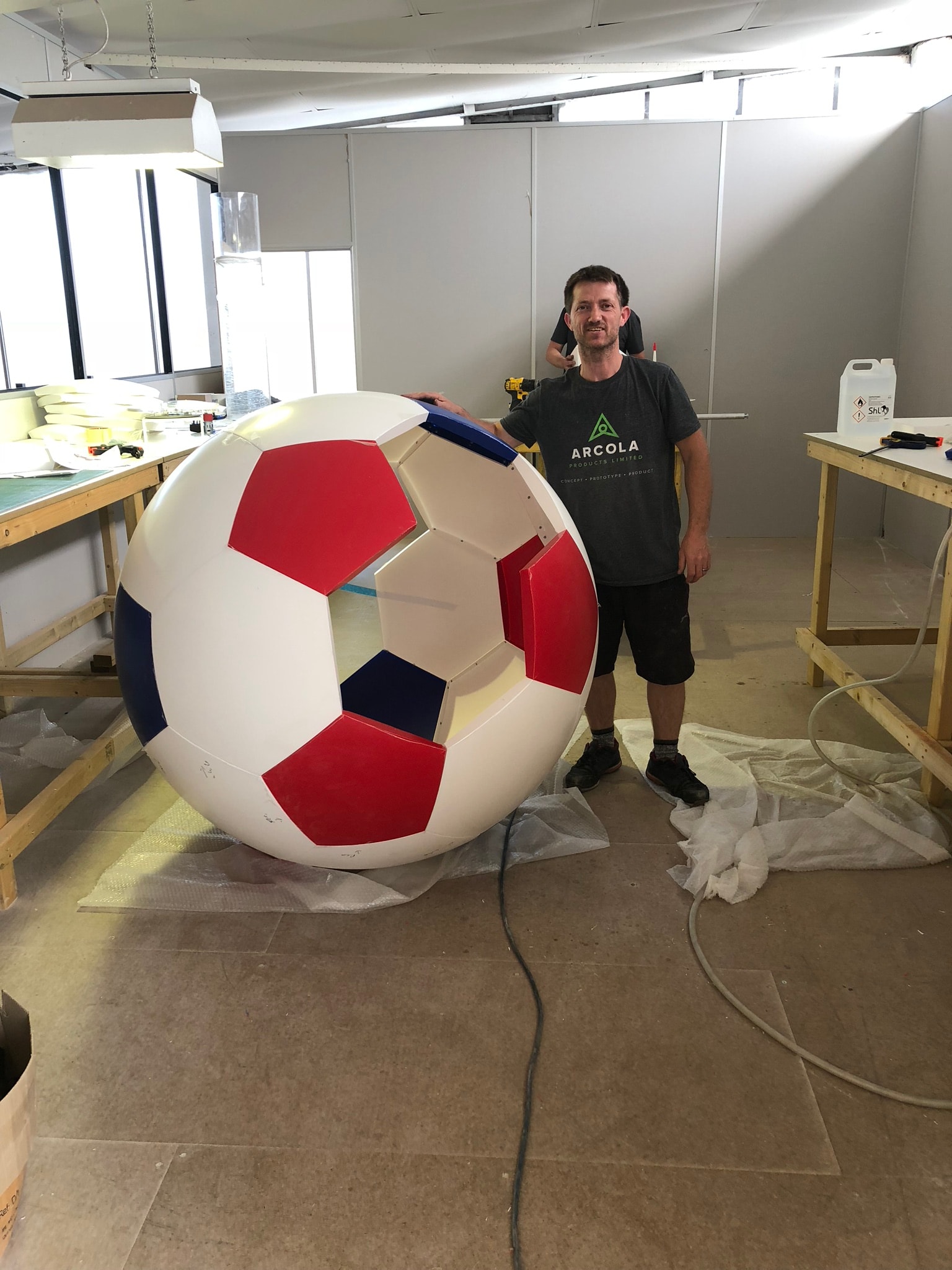 Giant vac-formed football shell with red and blue panels in a fabrication workshop, shown with a technician for scale; example of a plastic display/prop in production.