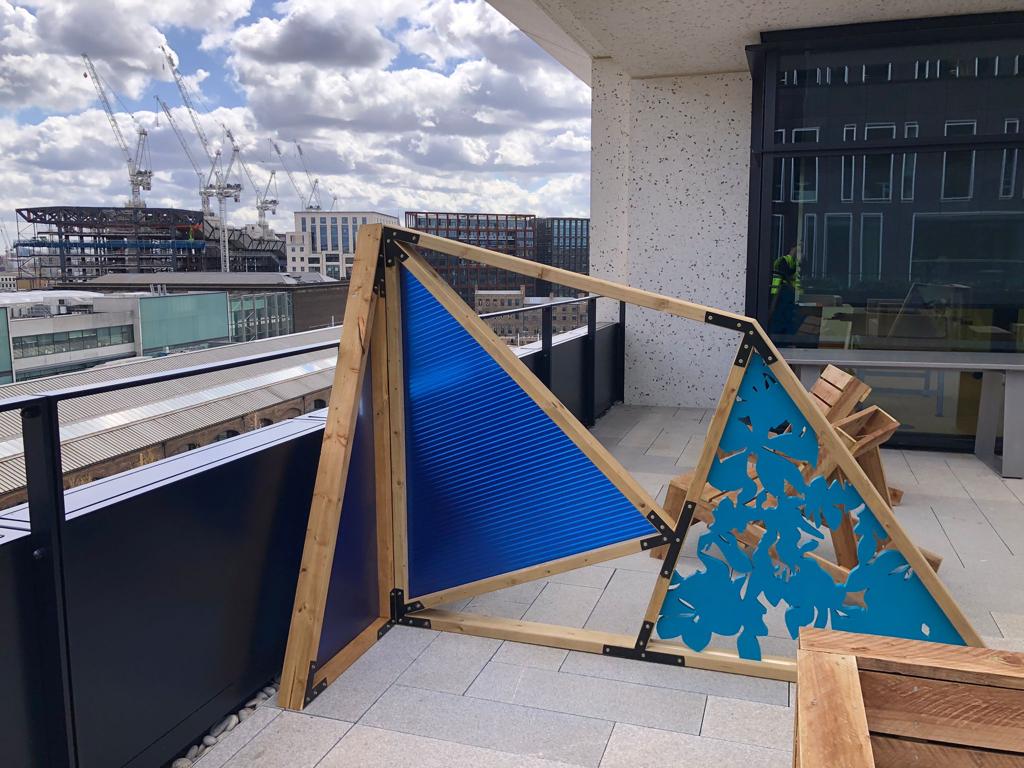 Finished triangular wooden and blue plastic decorative screen installed on a rooftop terrace, featuring a corrugated panel and a laser-cut decorative panel.