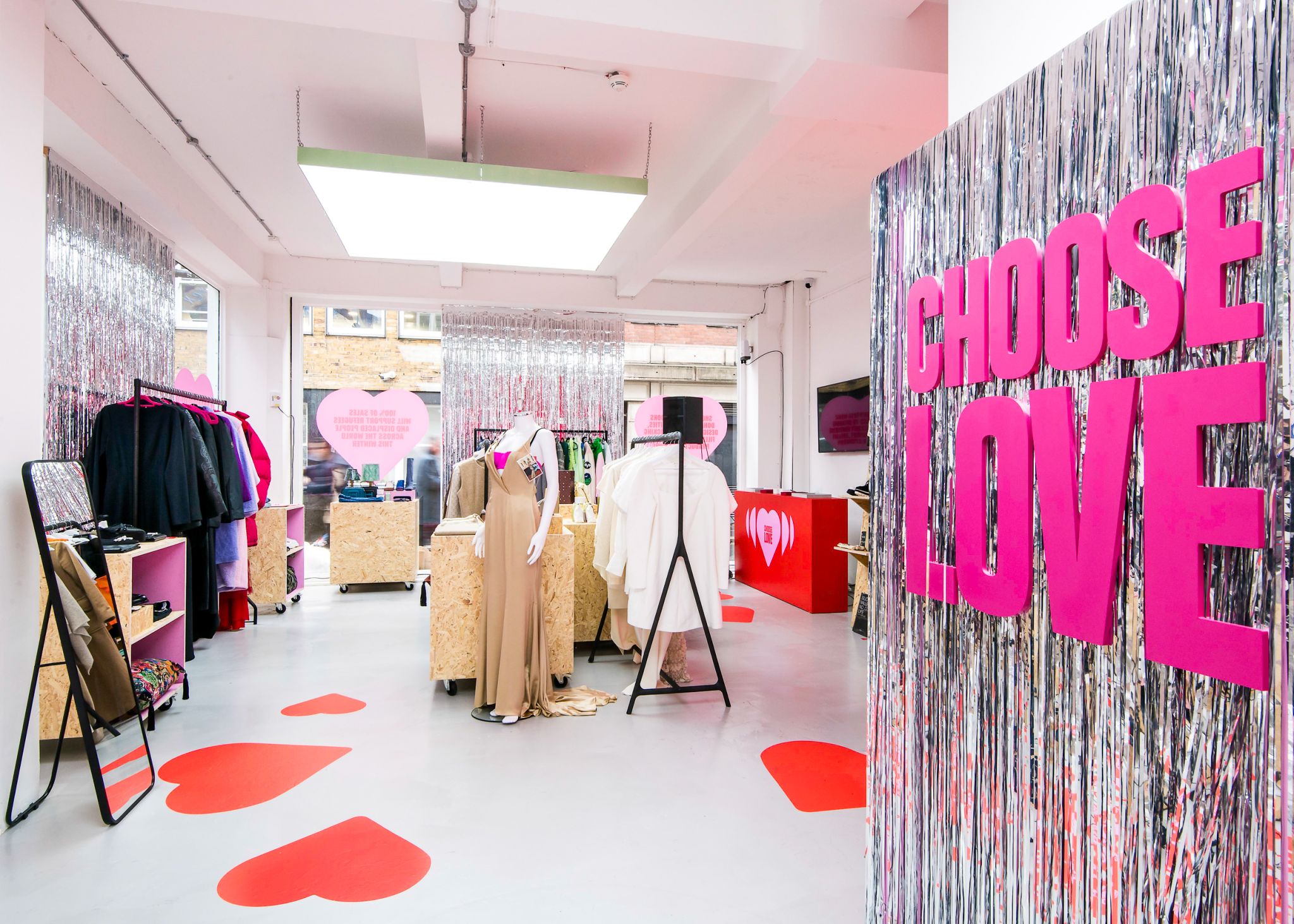 Bright pop-up shop interior with bold pink 'CHOOSE LOVE' wall lettering, clothing displays, mannequins and heart graphics — a branded temporary retail installation.