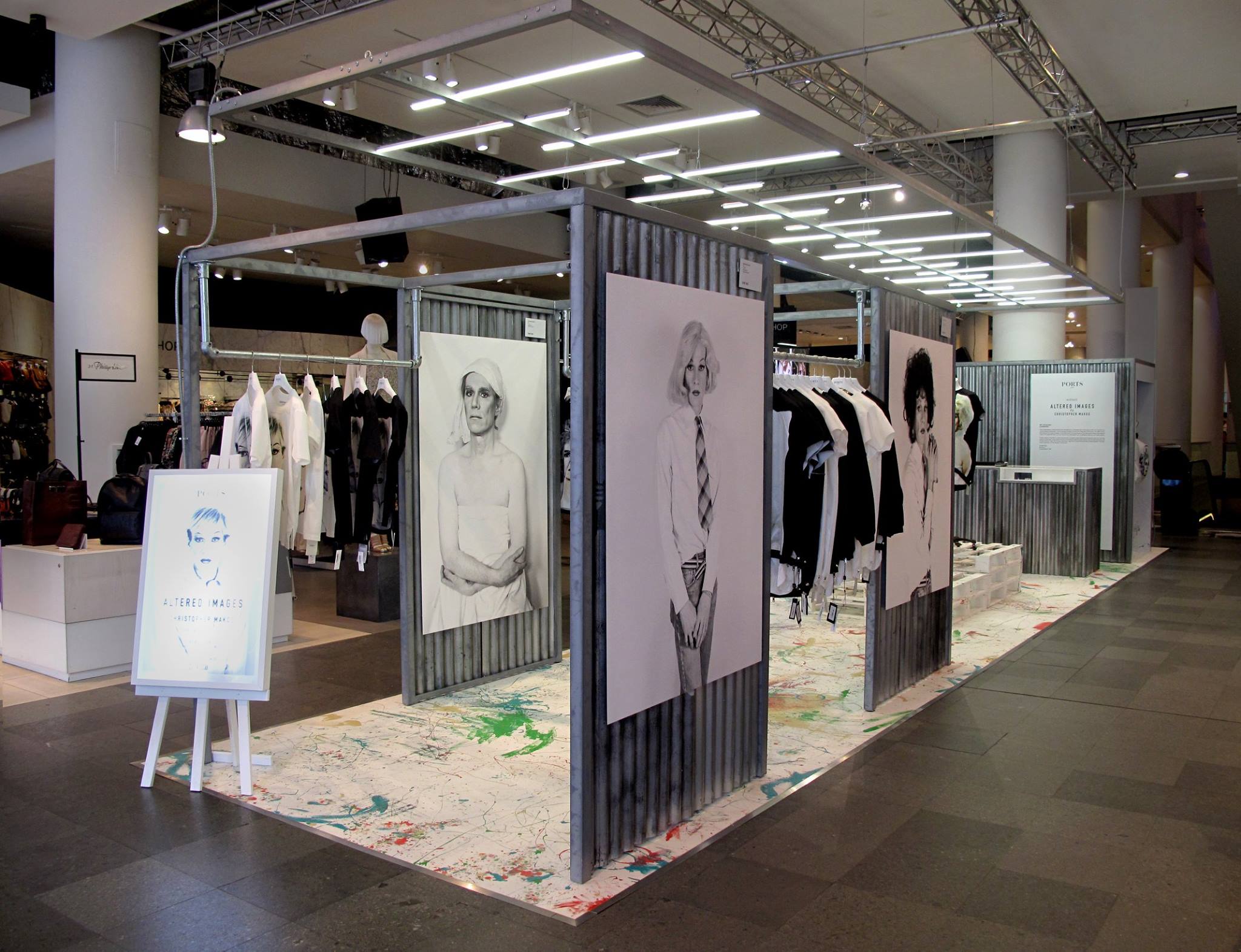Temporary pop-up retail installation inside a department store featuring corrugated partition panels, large portrait prints, clothing racks and an illuminated overhead display grid.