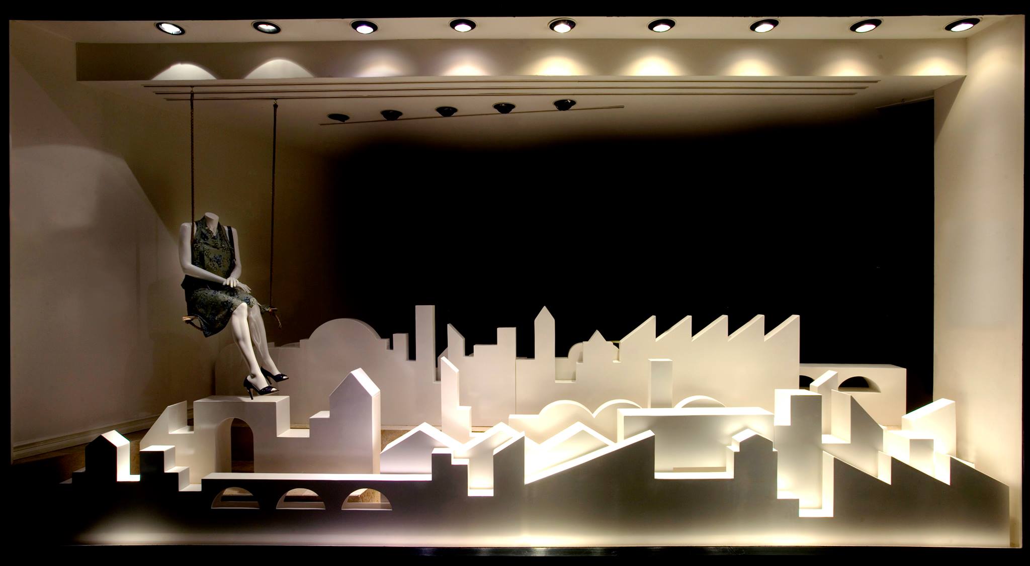 Dramatic shop window display with a suspended mannequin and backlit white architectural skyline models creating a striking retail presentation.