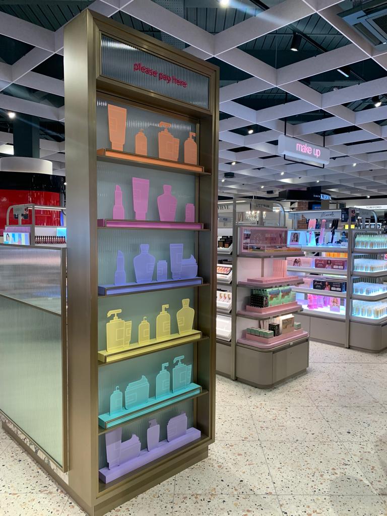Interior of a cosmetics retail department showing backlit display shelving with colorful product silhouette graphics and nearby product islands, plus directional signage for the makeup area.