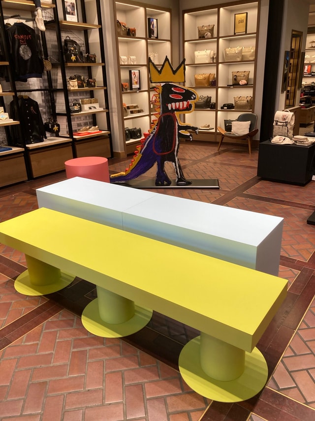 Retail store interior showing colorful display benches and a freestanding art installation in front of illuminated handbag shelving, suitable for store merchandising and fixture photography.
