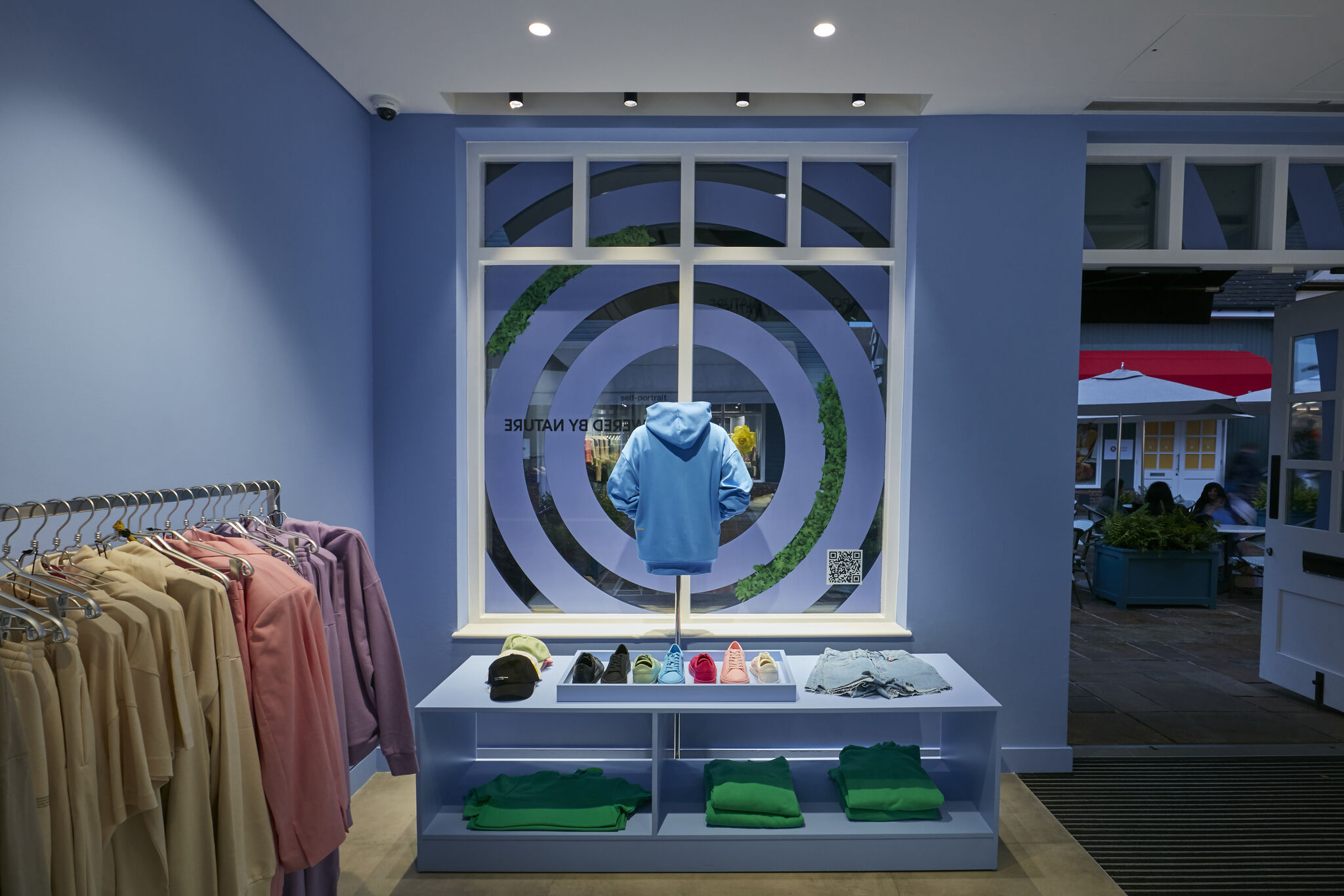 Well-lit retail shop interior featuring clothing racks, a central display table with folded garments and shoes, and a prominent window vinyl graphic behind a displayed hoodie.