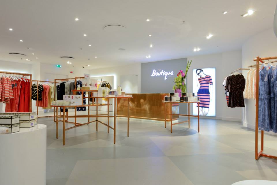 Bright modern boutique interior with clothing racks, central display tables and illuminated brand signage, featuring a backlit advertising panel and polished checkout counter.