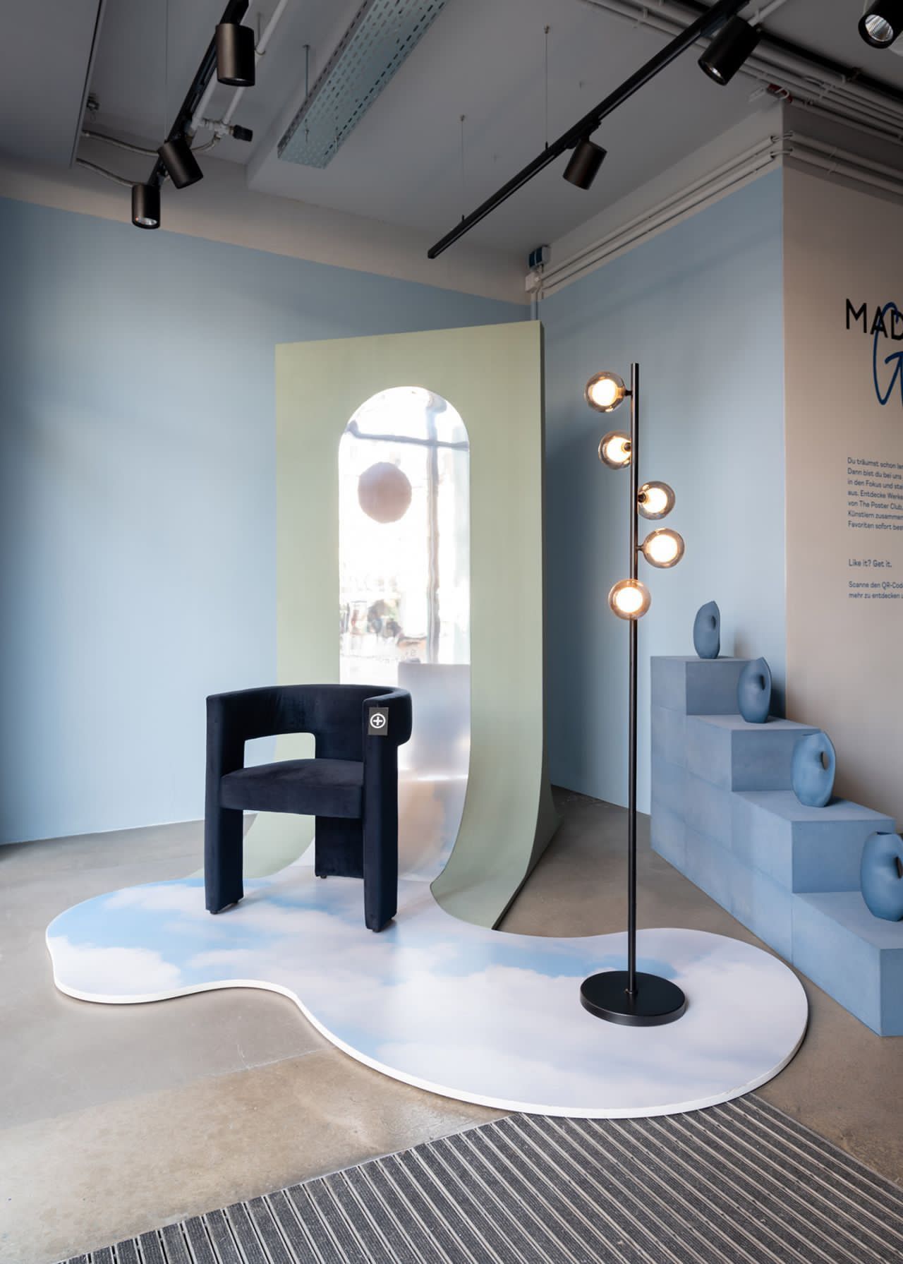 Styled retail showroom vignette featuring a sculptural chair on a cloud-printed display platform with an arched backdrop and decorative plinths, suitable for merchandising or brand display.