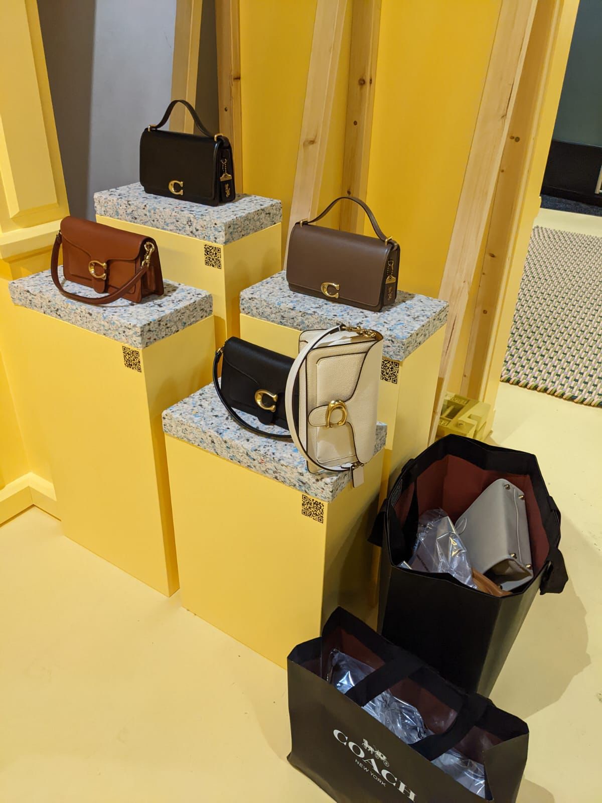 Coach handbags displayed on yellow pedestal plinths with terrazzo tops in a retail fit-out, featuring branded shopping bags and QR codes for product information.