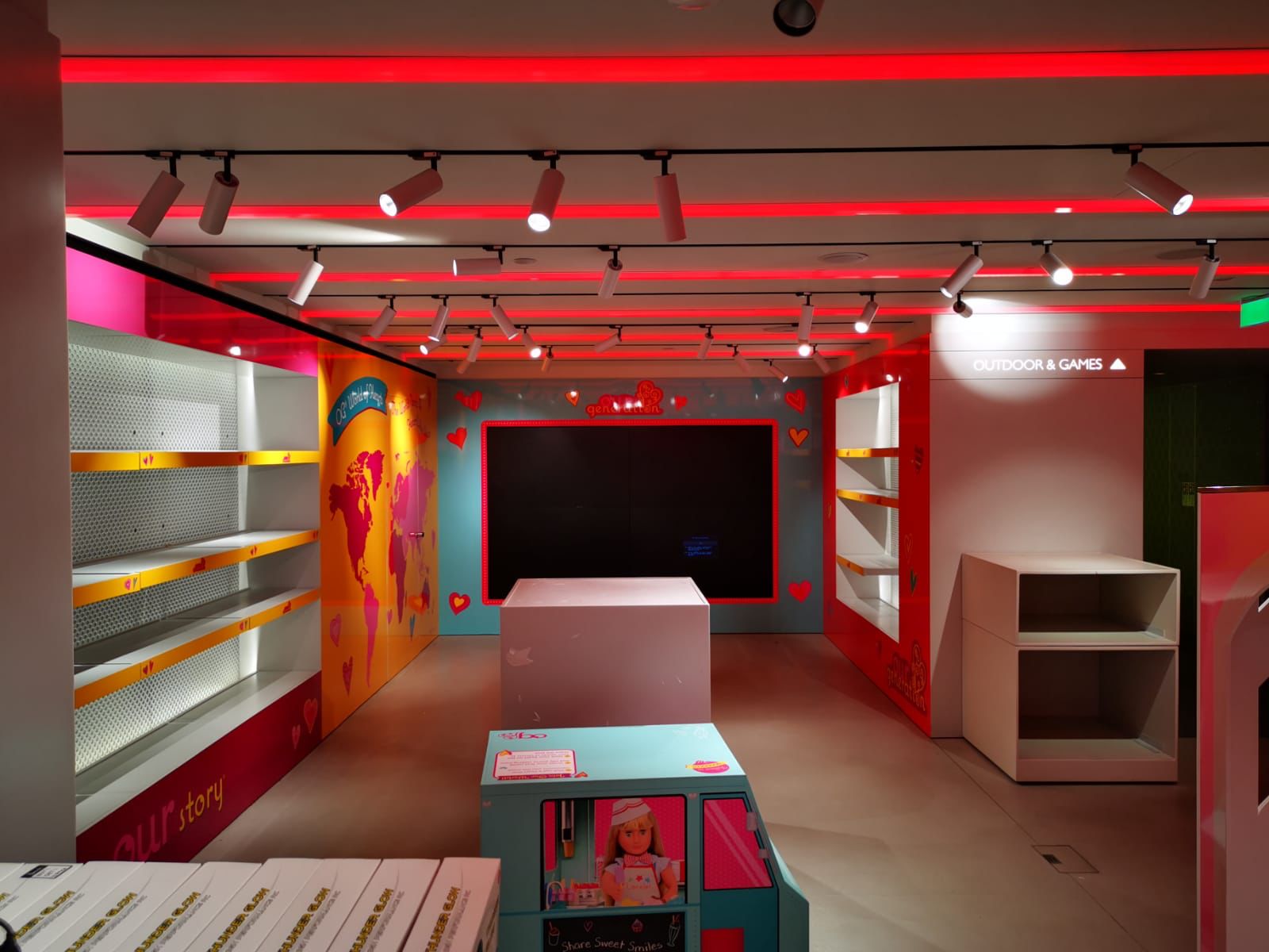 Finished retail interior with colourful printed wall graphics, shelving and display plinths under red accent lighting, featuring a section sign for 'Outdoor & Games'.