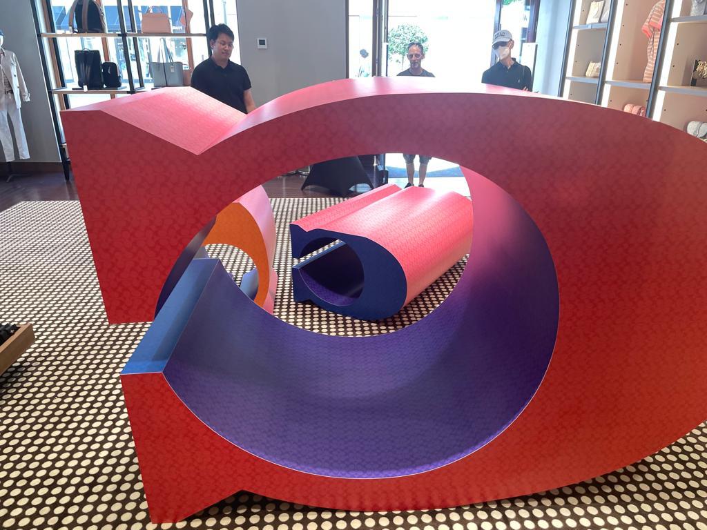 Large colorful sculptural display fixtures installed in a boutique retail store, photographed on a patterned floor with shelving and handbags in the background.