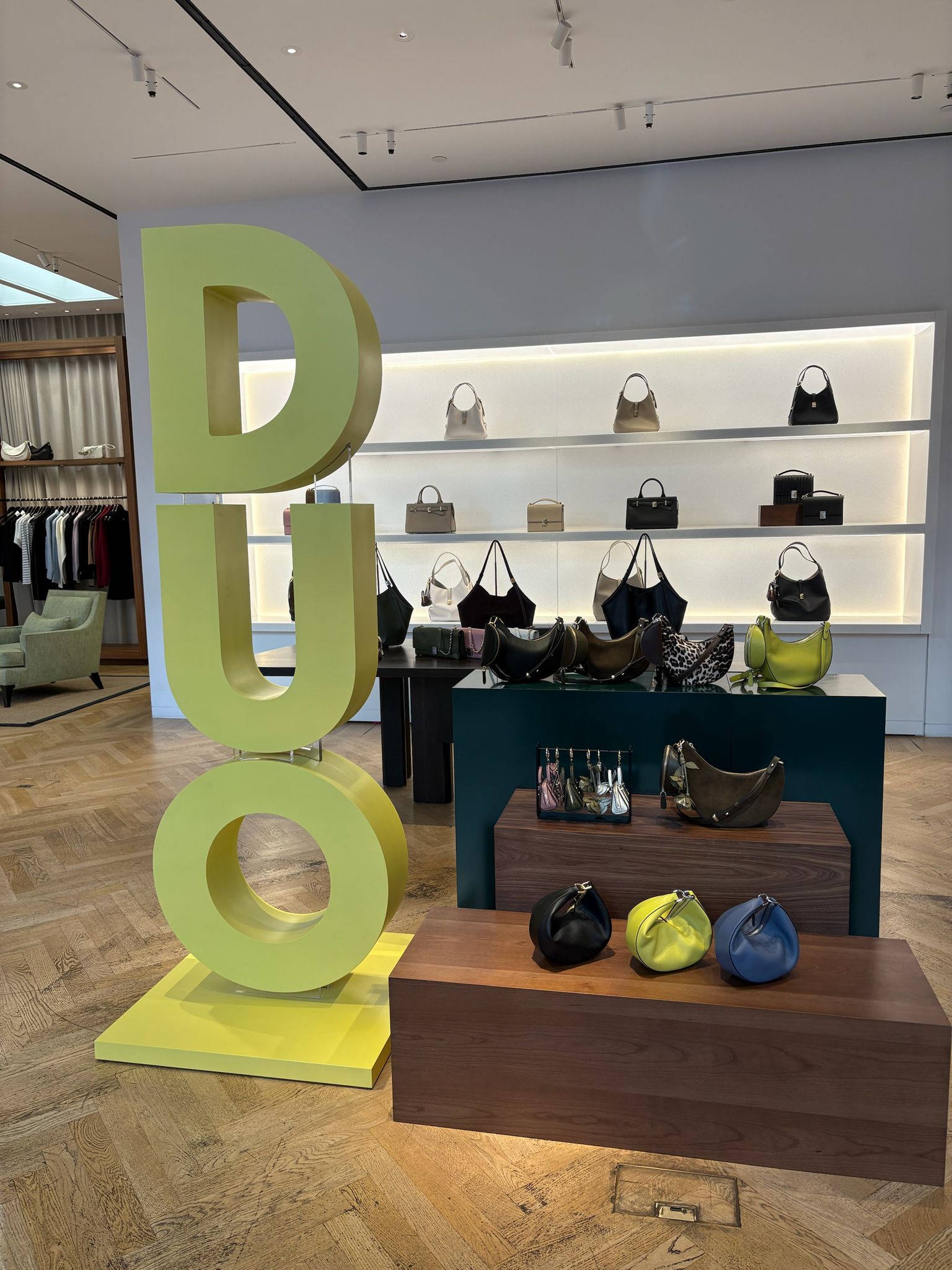 High-end retail interior showing a display of handbags on wooden plinths and backlit shelves with a large freestanding yellow 'DUO' sculptural prop for visual merchandising.