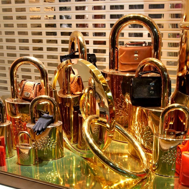 Luxury retail window display featuring oversized gold padlock sculptures and designer handbags on a reflective surface, creating a high-end visual merchandising installation.