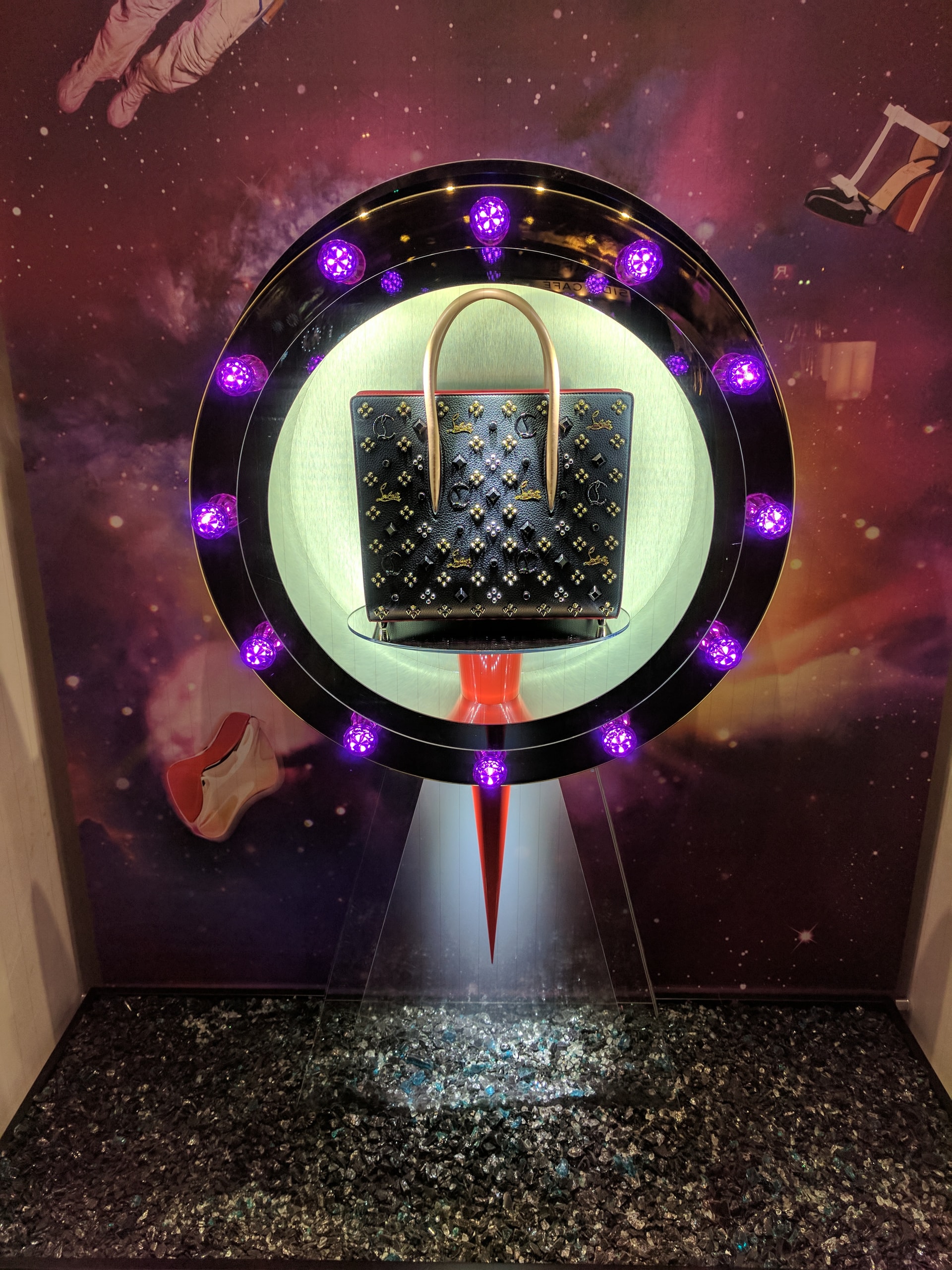 Retail window display featuring a studded designer handbag in a circular illuminated fixture with purple LED lights and a printed space-themed backdrop.